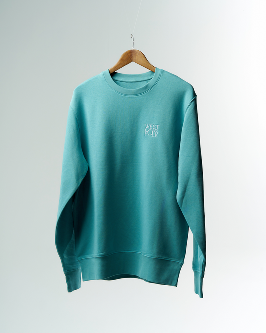 A turquoise West Cork Folk sweater with the logo embroidered on the chest, hanging on a wooden hanger against a white background.